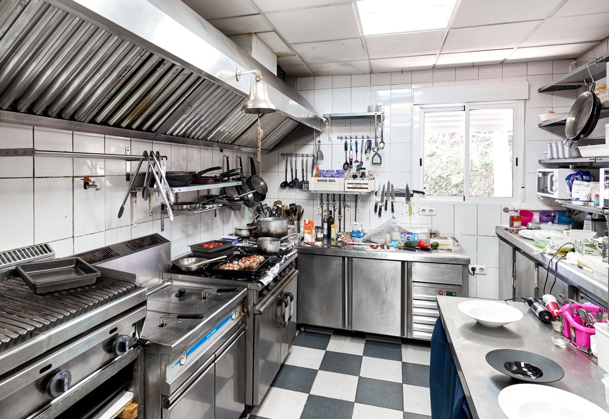 Commercial kitchen with stainless steel benches, a cooking range under an extraction hood and a black and white chequered floor