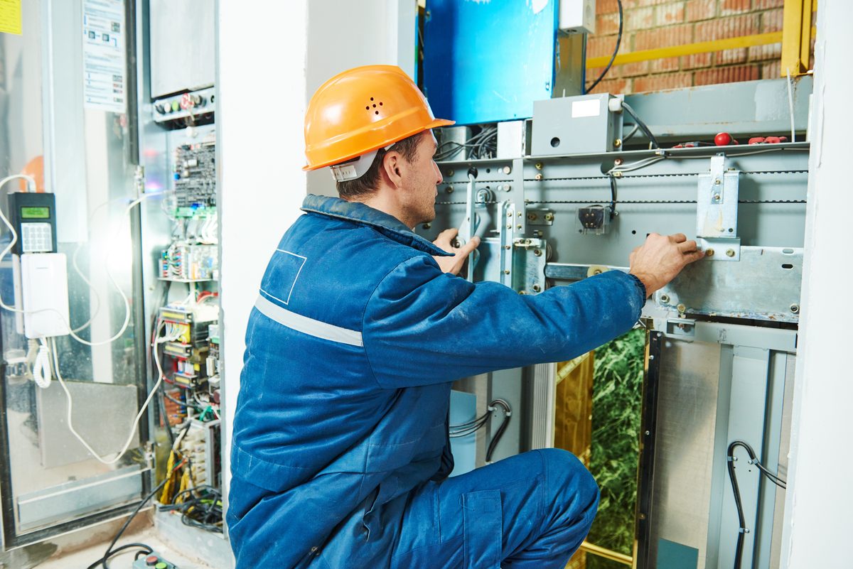 Electrician in blue overalls and an orange hard hat working inside a large grey electrical control panel