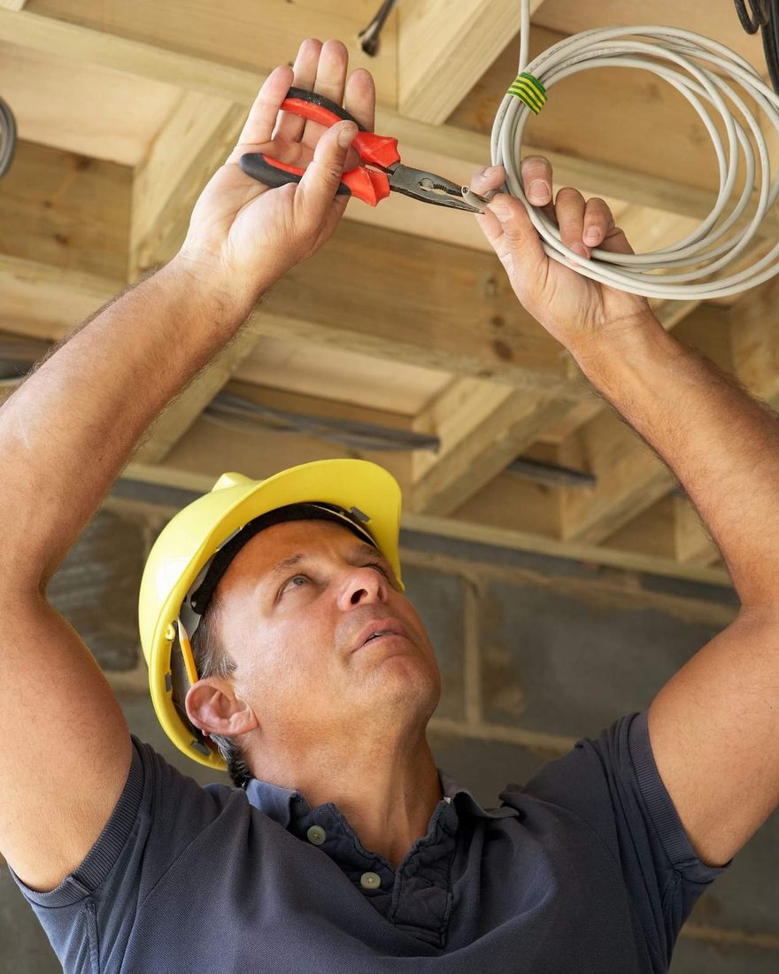 Electrician in a yellow hard hat cutting a cable among exposed timber ceiling framing, a coil of cable over one arm