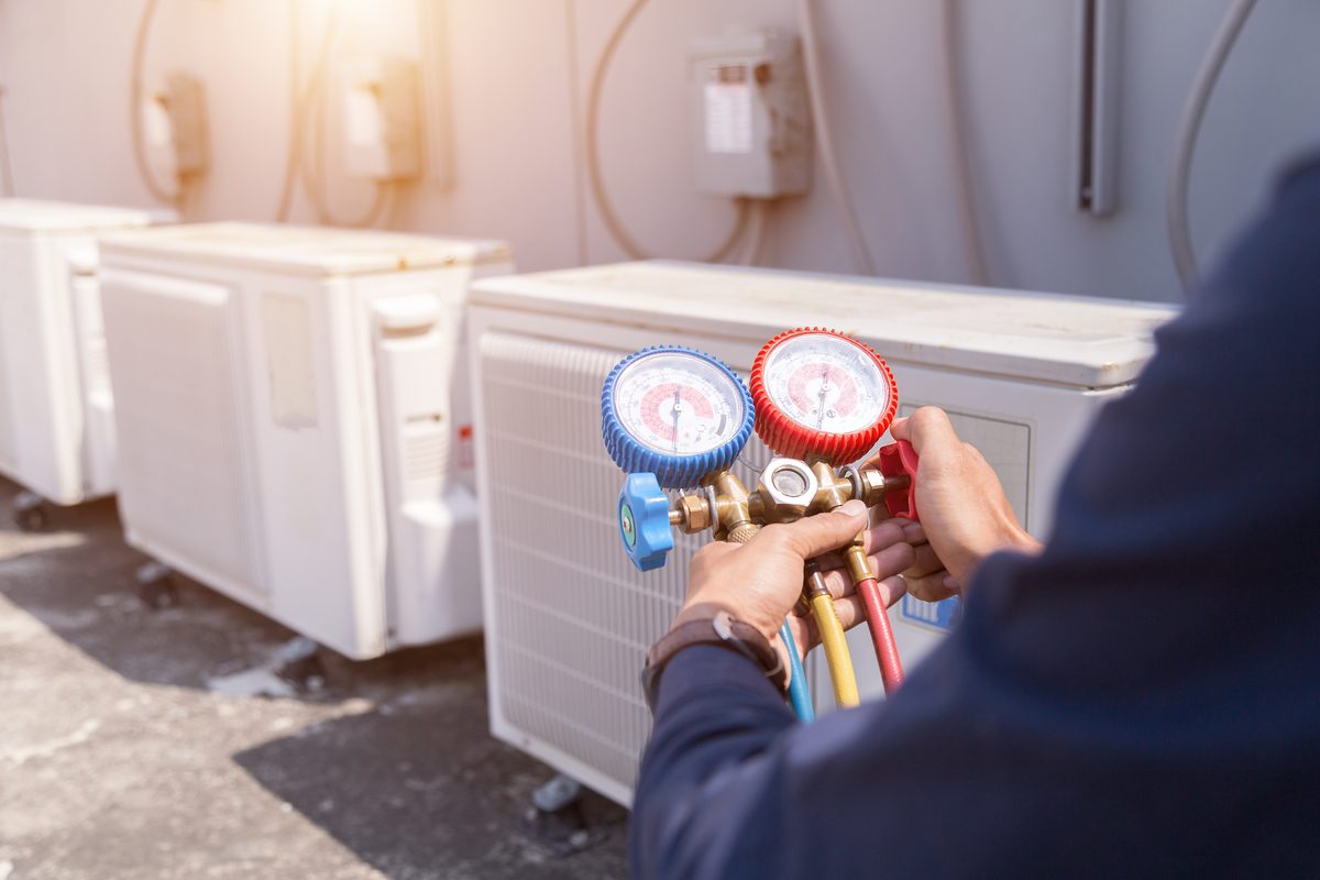 Technician holding red and blue refrigerant gauges beside a row of white heat pump outdoor units