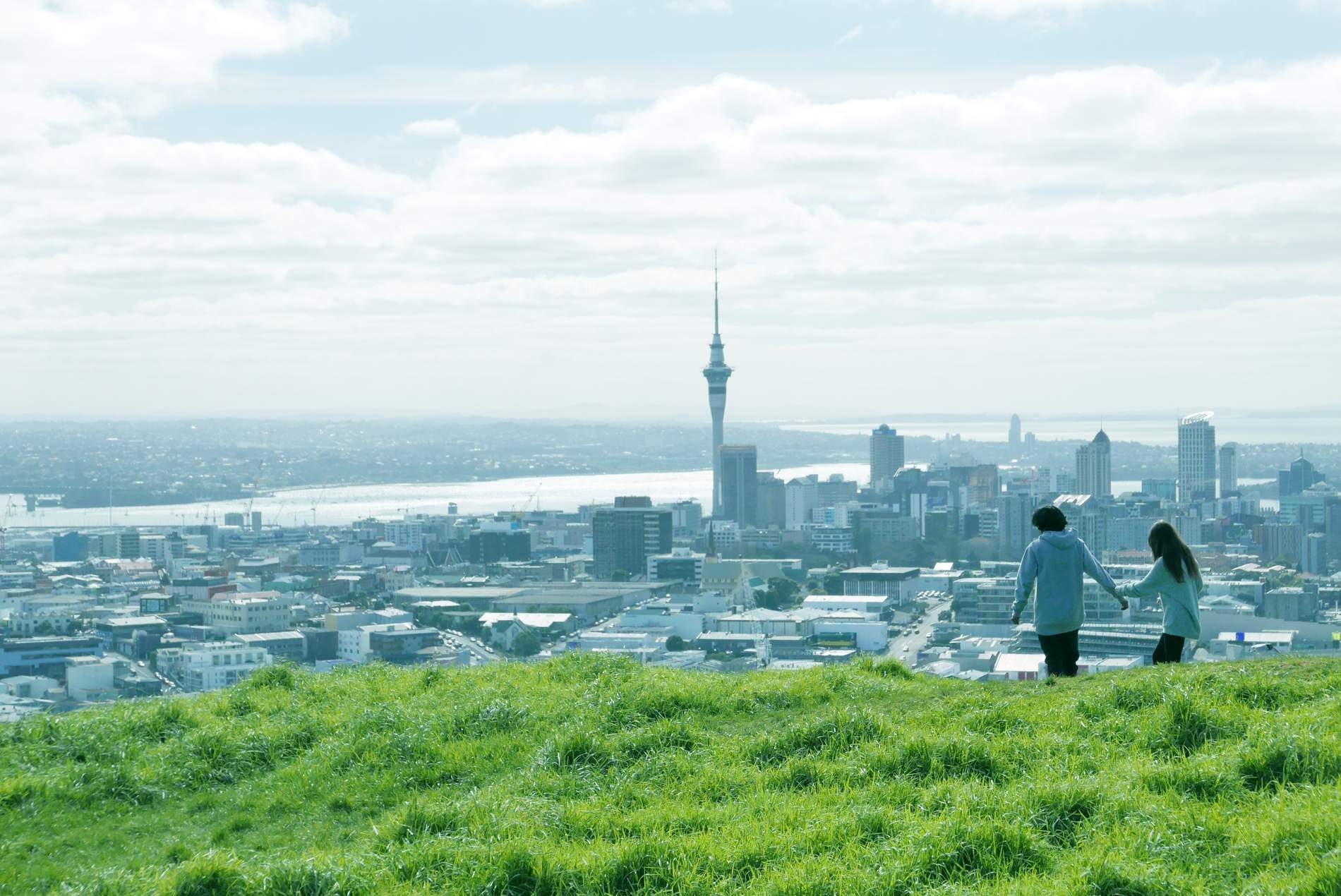 Auckland's city centre and the Sky Tower seen from the grassy summit of Mount Eden, the harbour beyond and two people walking along the slope