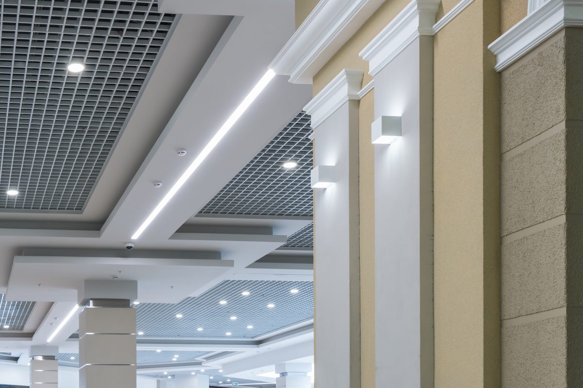 Commercial ceiling with grid panels, round recessed downlights and a long LED strip beside pale columns